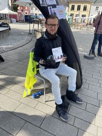 Alltags-Fitness-Test Ein junger Mann, sitzend auf einem Stuhl, mit einer Urkunde in der Hand. An Armen, Beinen, Oberkörper und Nacken trägt er Gewichtsmanschetten.