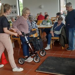 Eine jüngere Frau erklärt einer älteren Dame mit Rollator, wie sie über eine vor ihr auf dem Boden liegende Platte kommt. Im Hintergrund stehen und sitzen weitere ältere Personen und schauen zu.