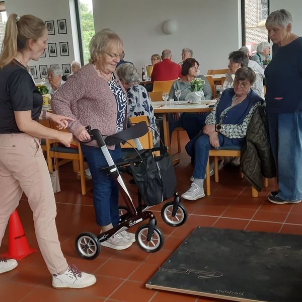 Eine jüngere Frau erklärt einer älteren Dame, wie sie mit dem Rollator über eine vor ihr auf dem Boden liegende Platte kommt. Im Hintergrund stehen und sitzen weitere ältere Menschen und schauen zu.