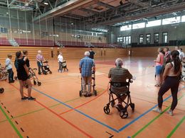Sicher unterwegs mit dem Rollstuhl oder Rollator Ein Mann im schwarzen T-Shirt steh in der Mitte einer Turnhalle. Mehrere Menschen stehen mit Rollatoren um ihn herum und hören zu.