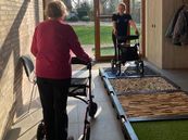 Zwei Personen mit Rollatoren in einem hellen Flur, eine Person steht auf einem taktilen Übungsparcours mit verschiedenen Oberflächen, die andere nähert sich von hinten.