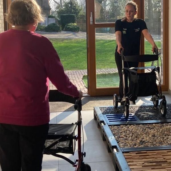 Zwei Personen mit Rollatoren in einem hellen Flur, eine Person steht auf einem taktilen Übungsparcours mit verschiedenen Oberflächen, die andere nähert sich von hinten.