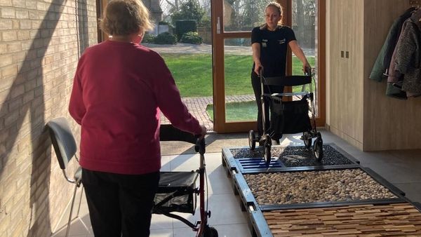 Zwei Personen mit Rollatoren in einem hellen Flur, eine Person steht auf einem taktilen Übungsparcours mit verschiedenen Oberflächen, die andere nähert sich von hinten.