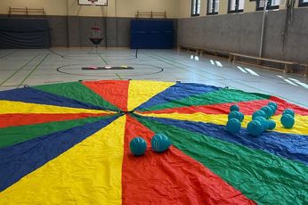 Schwungtuch und Discgolf stehen bereit. Ein großes, buntes Schwungtuch liegt ausgebreitet in einer Turnhalle. Auf ihr liegen knapp zwei Duzend türkise Luftballons. Im Hintergrund ist das Spiel Discgolf zu sehen.