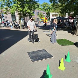 Zwei Personen stehen auf einem gepflasterten Platz mit Bäumen und Gebäuden. Um sie herum ist ein Rollatorparcours aufgebaut.