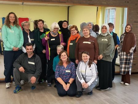 Gruppe von Menschen unterschiedlichen Alters in einem hellen Raum mit Holzfußboden und gelben Wänden, einige sitzen, andere stehen.