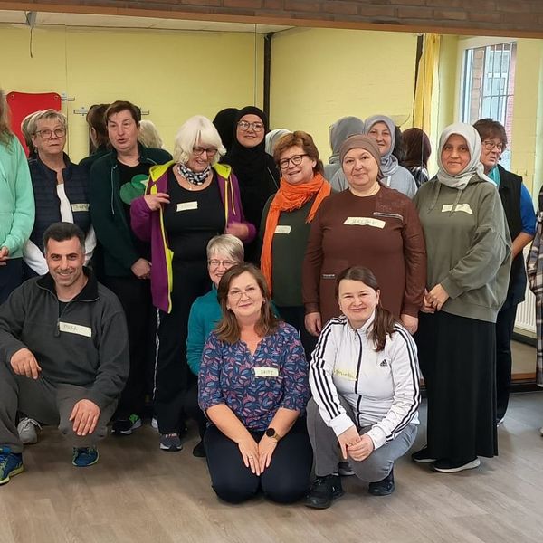 Gruppe von Menschen unterschiedlichen Alters in einem hellen Raum mit Holzfußboden und gelben Wänden, einige sitzen, andere stehen.