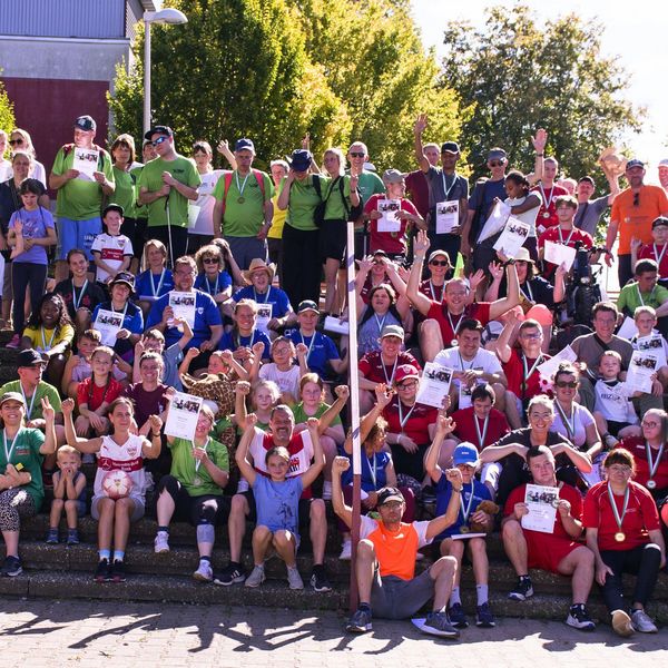 Große Gruppe von Menschen unterschiedlichen Alters und Geschlechts sitzt und steht auf Treppenstufen im Freien, viele halten Urkunden hoch und tragen Medaillen.