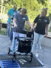 Auch die Mitarbeitenden werden geschult. Eine Pflegekraft in grauer Kleidung benutzt einen Rollator, während sie eine Rampe hinuntergeht. Im Hintergrund sind weitere Personen sichtbar.