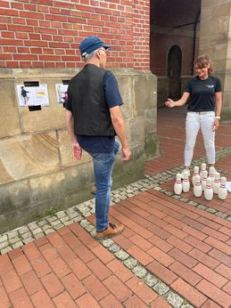 Ein älterer Mann mit einer Cappy steht mit dem Rücken zur Kamera und hebt sein Knie an. Eine Mitarbeiterin des Kreissportbundes steht vor ihm und zählt mit Hilfe eines Zählers seine Wiederholungen.