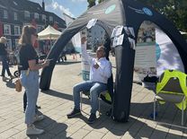 Alltags-Fitness-Test Ein Mann im weißen Hemd und blauer Jeans, sitzend auf einem Stuhl unter dem Zelt des Kreissportbundes, der mit den Händen zeigt, wie hoch etwas ist. Eine Mitarbeiterin des Kreissportbundes steht vor ihm und hört zu.