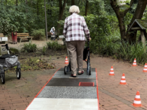 Auf dem Weg zum Rollator-Führerschein Eine ältere Dame geht mit dem Rollator von der Kamera weg über ein Hindernis aus Kopfsteinpflaster, Fliesen und Rasen. Neben ihr stehen mehrere Pylonen.