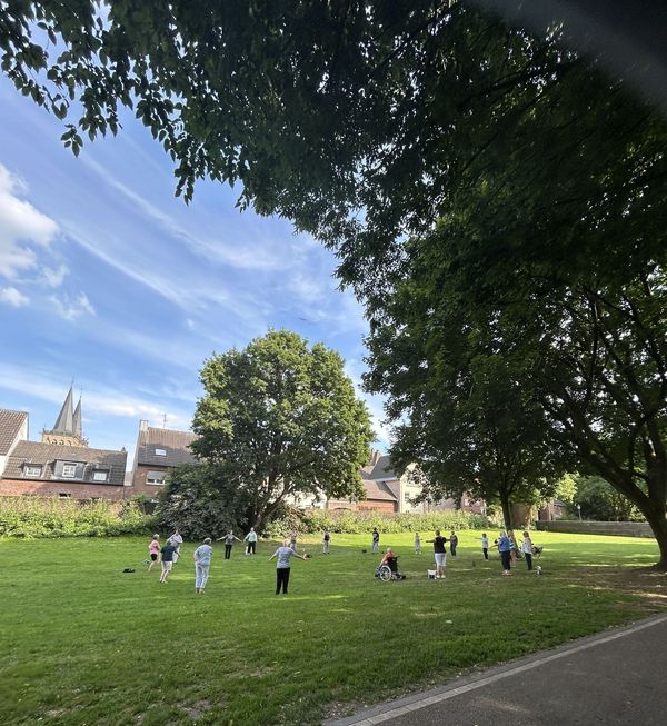 Gruppe von Menschen unterschiedlichen Alters steht und sitzt auf einer Wiese unter großen Bäumen in einem Park, im Hintergrund Häuser und Kirchturm.