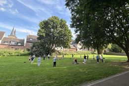 Gruppe von Menschen unterschiedlichen Alters steht und sitzt auf einer Wiese unter großen Bäumen in einem Park, im Hintergrund Häuser und Kirchturm.