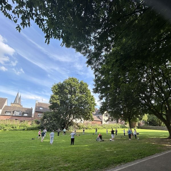 Gruppe von Menschen unterschiedlichen Alters steht und sitzt auf einer Wiese unter großen Bäumen in einem Park, im Hintergrund Häuser und Kirchturm.