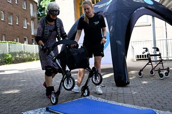 Im Alterungsanzug durch den Parcours. Eine Frau im gemusterten Kleid, mit Gewichtsmanschetten an Ellbogen, Hand-, Knie- und Fußgelenken kippt einen Rollator zum Überqueren einer blauen Turnmatte. Neben ihr steht eine Mitarbeiterin des Kreissportbundes und gibt ihr Tipps.