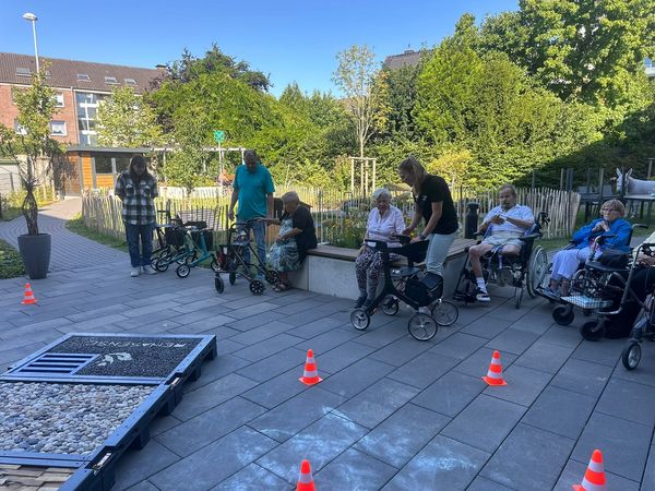 Personen mit Rollatoren stehen auf einem gepflasterten Weg. Im Hintergrund sind Bäume und Sträucher sichtbar. Rote Kegel sind auf dem Boden verteilt.