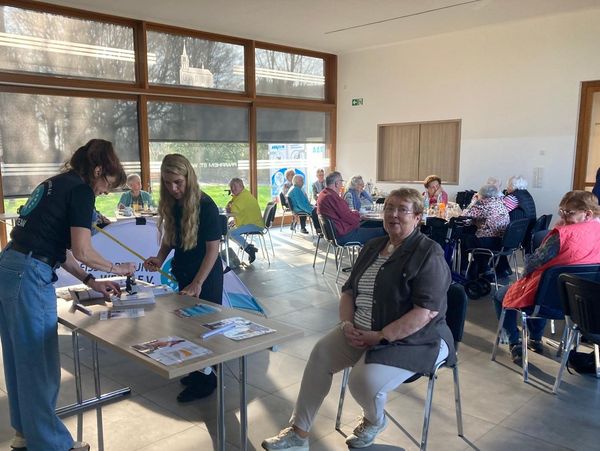 Mehrere ältere Menschen sitzen an Tischen in einem hellen Raum mit großen Fenstern, zwei jüngere Personen stehen an einem Tisch mit Flyern und Broschüren.