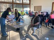 Mehrere ältere Menschen sitzen an Tischen in einem hellen Raum mit großen Fenstern, zwei jüngere Personen stehen an einem Tisch mit Flyern und Broschüren.