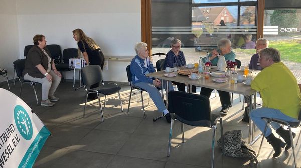 Mehrere ältere Personen sitzen an einem Tisch mit Getränken und Snacks in einem hellen Raum mit großen Fenstern.