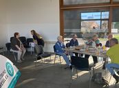 Mehrere ältere Personen sitzen an einem Tisch mit Getränken und Snacks in einem hellen Raum mit großen Fenstern.