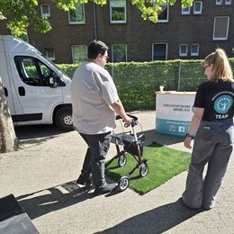 Mann mit Rollator geht auf einem Stück Kunstrasen neben einer Frau in Team-T-Shirt.