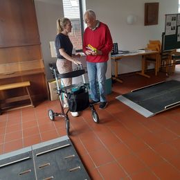 Eine jüngere Frau überreicht einem älteren Herrn mit Rollator Präsente. Im Vordergrund Platten eines Rollatorparcours.