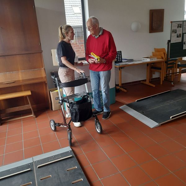 Eine jüngere Frau überreicht einem älteren Herrn Präsente. Im Vordergrund liegen Platten eines Rollatorparcours.