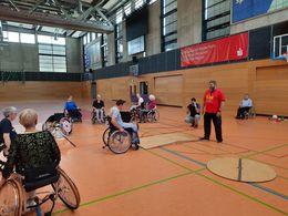 Sicher unterwegs mit dem Rollstuhl oder Rollator Ein Mann in einem roten T-Shirt steht vor einer Holzrampe. Mehrere Menschen, in Rollstühlen sitzend, fahren um die Rampe.