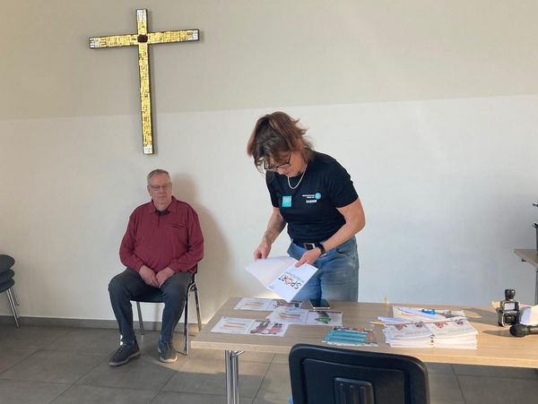Mann sitzt auf Stuhl vor Wand mit großem Kreuz, Frau steht an Tisch mit Papieren und Ordnern in hellem Raum.