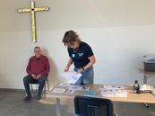 Mann sitzt auf Stuhl vor Wand mit großem Kreuz, Frau steht an Tisch mit Papieren und Ordnern in hellem Raum.