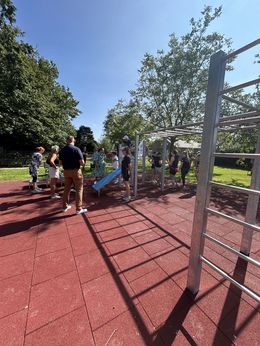 Einige Menschen stehen bei Sonnenschein zusammen vor einem Gerät eines Outdoorparcours.