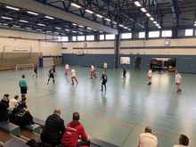 Hallensportspiel mit zwei Mannschaften in rot-weißer und dunkelblauer Sportkleidung auf Spielfeld mit Toren und Zuschauern auf der Tribüne.