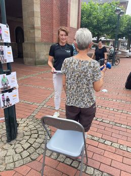 Eine ältere Dame steht mit dem Rücken zu einem Stuhl und macht im Stehen mit einer Hantel eine Übung für den Bizeps. Eine Mitarbeiterin des Kreissportbundes steht vor ihr und gibt ihr Tipps.