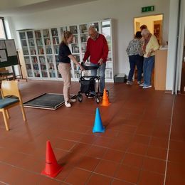 Eine jüngere Frau erklärt einem älteren Herrn einen Rollator. Im Vordergrund stehen Pylonen eines Rollatorparcours.