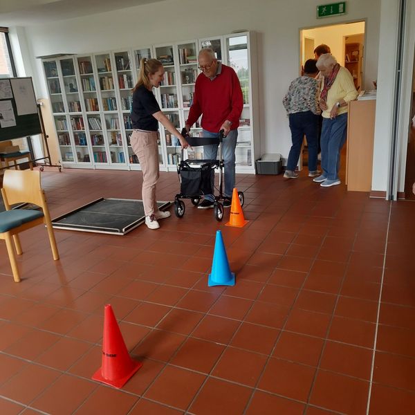 Eine jüngere Frau erklärt einem älteren Herrn einen Rollator. Im Vordergrund stehen Pylonen eines Rollatorparcours.