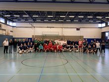 Gruppenfoto von mehreren Fußballmannschaften, Schiedsrichtern und Ehrengästen in einer Sporthalle vor einem Handballtor.