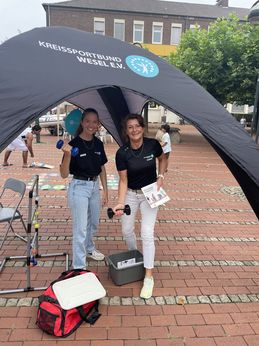 Zwei Mitarbeiterinnen des Kreissportbundes in schwarzen T-Shirts stehen unter einem Pavillon mit Hanteln in der Hand.
