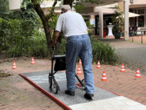 Auf dem Weg zum Rollator-Führerschein Ein älterer Herr geht mit dem Rollator von der Kamera weg über ein Hindernis aus Kopfsteinpflaster, Fliesen und Rasen. Neben ihm stehen mehrere Pylonen.
