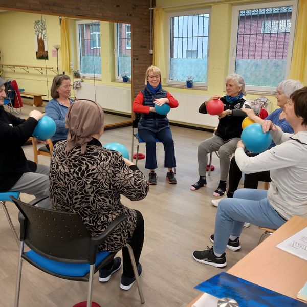 Gruppe von Erwachsenen sitzt im Stuhlkreis in einem hellen Raum und hält bunte Bälle in den Händen.