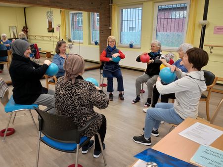 Gruppe von Erwachsenen sitzt im Stuhlkreis in einem hellen Raum und hält bunte Bälle in den Händen.