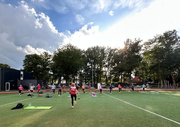 Mehrere Frauen stehen mit Abstand auf einer Wiese, einige auf Yogamatten, bei sonnigem Himmel mit Wolken und Bäumen im Hintergrund.