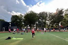 Mehrere Frauen stehen mit Abstand auf einer Wiese, einige auf Yogamatten, bei sonnigem Himmel mit Wolken und Bäumen im Hintergrund.