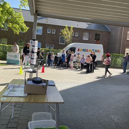 Menschen, darunter Kinder, stehen vor einem weißen Van mit der Aufschrift 'SPIELMOBIL' auf einem gepflasterten Platz neben einem Gebäude.