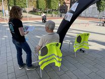 Alltags-Fitness-Test Ein älterer Mann mit dem Rücken zur Kamera, sitzend auf einem Stuhl, während eine Mitarbeiterin des Kreissportbundes mit einem Flyer neben ihm steht und erklärt.