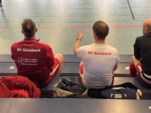 Drei Männer in Sportkleidung sitzen auf einer Bank in einer Halle, zwei tragen Trikots mit der Aufschrift 'SV Sonsbeck' auf dem Rücken, einer zeigt mit der rechten Hand nach vorne.