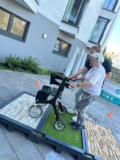 Übungen auf den verschiedenen Oberflächen des Parcours. Eine ältere Frau mit einem Rollator steht auf einer Plattform mit unterschiedlichen Oberflächen. Eine Mitarbeiterin des KSB hilft ihr dabei. Im Hintergrund sind Gebäude und Pflanzen sichtbar.