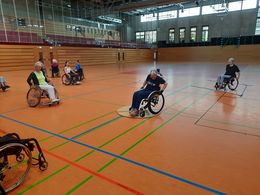 Mehrere Menschen, in Rollstühlen sitzend, fahren durch eine Turnhalle. Eine Rollstuhlfahrerin steht auf einem runden Holzbrett.