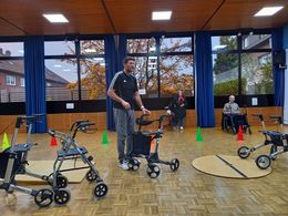 Aktionstag sicher unterwegs mit dem Rollstuhl und Rollator Ein Mann steht mit einem Rollator in einer Turnhalle und erklärt. Eine Frau und ein Mann stehen im Hintergrund und hören zu. Im Vordergrund stehen Rollatoren.