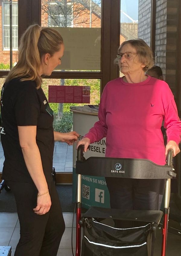 Eine ältere Frau mit Rollator spricht mit einer jüngeren Frau in schwarzer Kleidung vor großen Fenstern in einem hellen Raum.